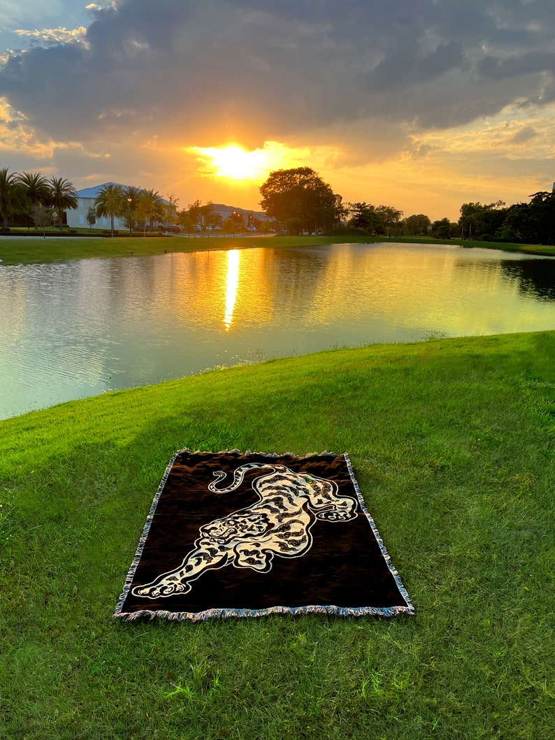May include: A brown and black woven throw blanket with a tiger design. The blanket is laid out on a grassy area near a lake with a sunset in the background.