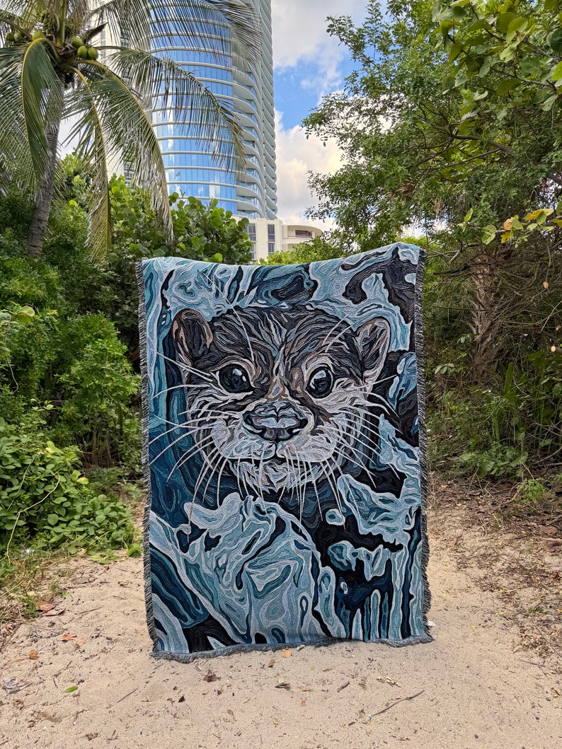 May include: A woven tapestry featuring a close-up of an otter's face with a blue and gray abstract background.