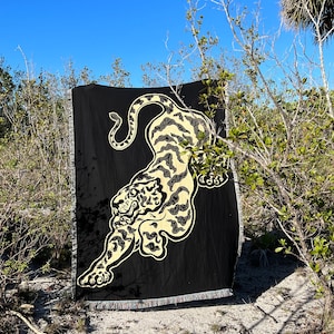 May include: A black woven throw blanket with a cream colored tiger design. The blanket is fringed and is laying on the ground in front of some bushes.