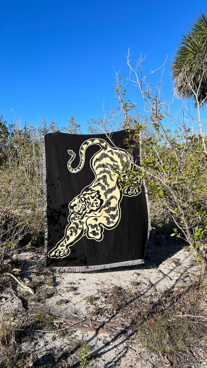 May include: A black woven throw blanket with a cream colored tiger design. The blanket is fringed and is laying on the ground in front of some bushes.