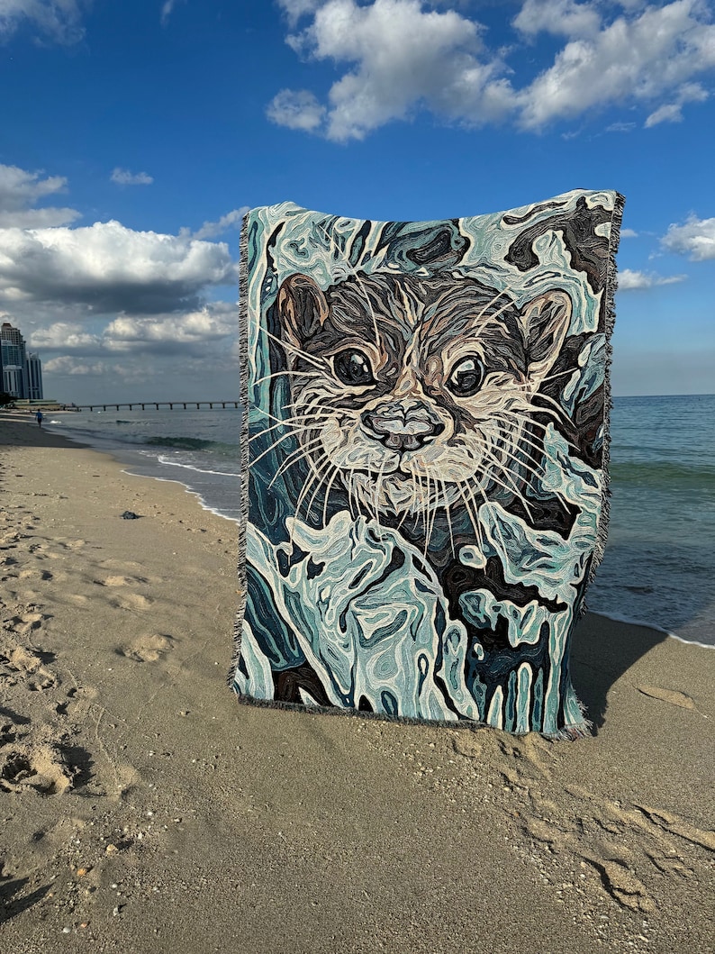 Peut inclure: Une tapisserie tiss&eacute;e repr&eacute;sentant un gros plan sur le visage d'une loutre sur un fond abstrait bleu et vert. La tapisserie est sur une plage de sable.