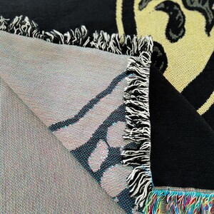 May include: A black and yellow woven throw blanket with a fringe border. The blanket features a graphic design of a face with a large nose and mouth.