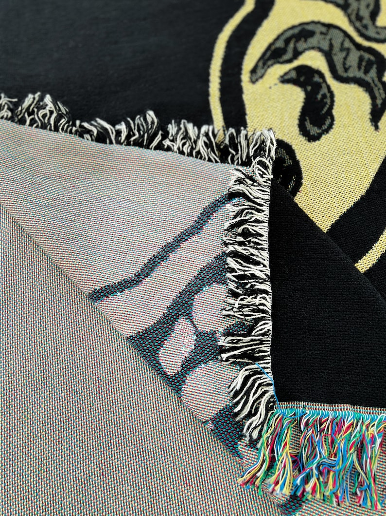 May include: A black and yellow woven throw blanket with a fringe border. The blanket features a graphic design of a face with a large nose and mouth.