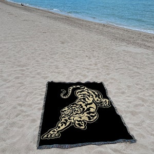 May include: A black woven throw blanket with a gold embroidered tiger design. The blanket is lying on a sandy beach.
