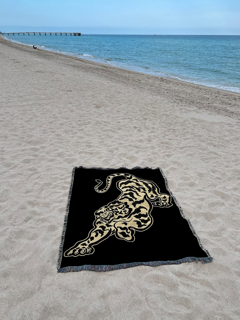 May include: A black woven throw blanket with a gold embroidered tiger design. The blanket is lying on a sandy beach.