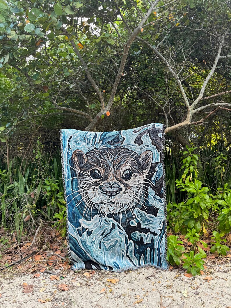 May include: A blue and white woven tapestry featuring a close-up of an otter's face. The otter's fur is brown and black, and its eyes are dark. The tapestry is hanging on a tree branch in a forest setting.