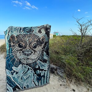 May include: A woven tapestry featuring a close-up of an otter's face. The tapestry is blue, green, and brown with a textured, abstract design. The tapestry is displayed on a sandy beach with a lifeguard stand in the background.