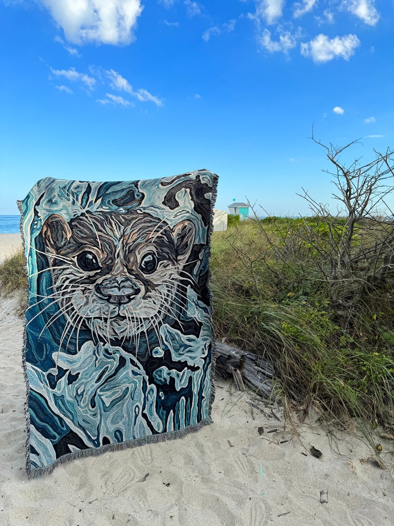 May include: A woven tapestry featuring a close-up of an otter's face. The tapestry is blue, green, and brown with a textured, abstract design. The tapestry is displayed on a sandy beach with a lifeguard stand in the background.