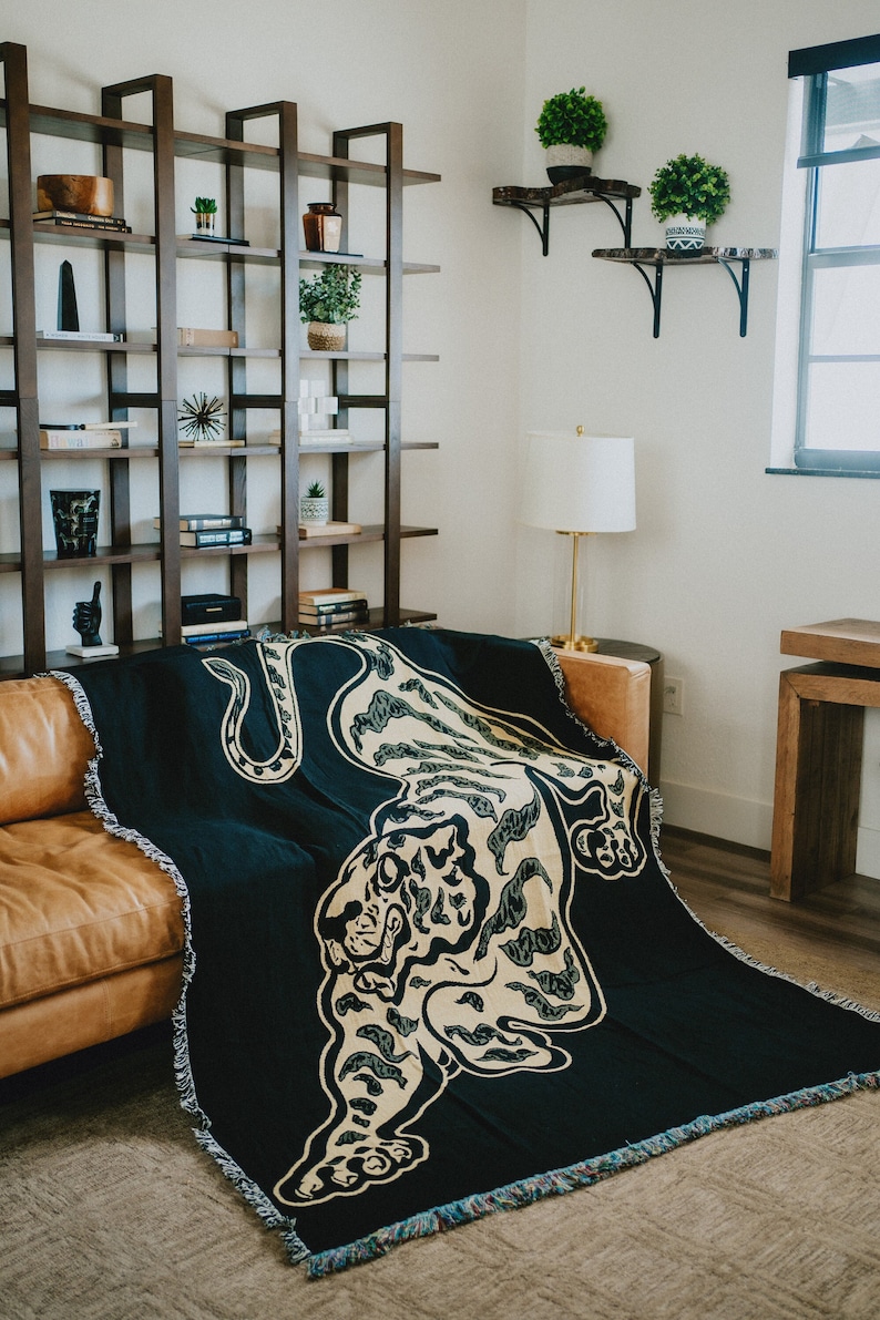 May include: A black and gold woven throw blanket with a tiger design. The blanket is draped over a brown leather couch in a living room setting.