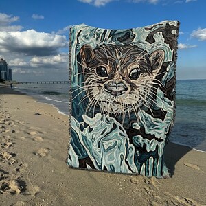 May include: A woven tapestry throw blanket featuring a close-up of an otter's face. The otter is depicted in shades of brown and white, with a blue and green background. The blanket is shown on a sandy beach.