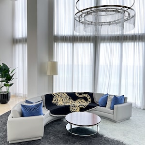 May include: A white curved sectional sofa with blue throw pillows and a black throw blanket with a gold leopard print. The sofa is in a modern living room with a large crystal chandelier and a glass-top coffee table with a wooden top.