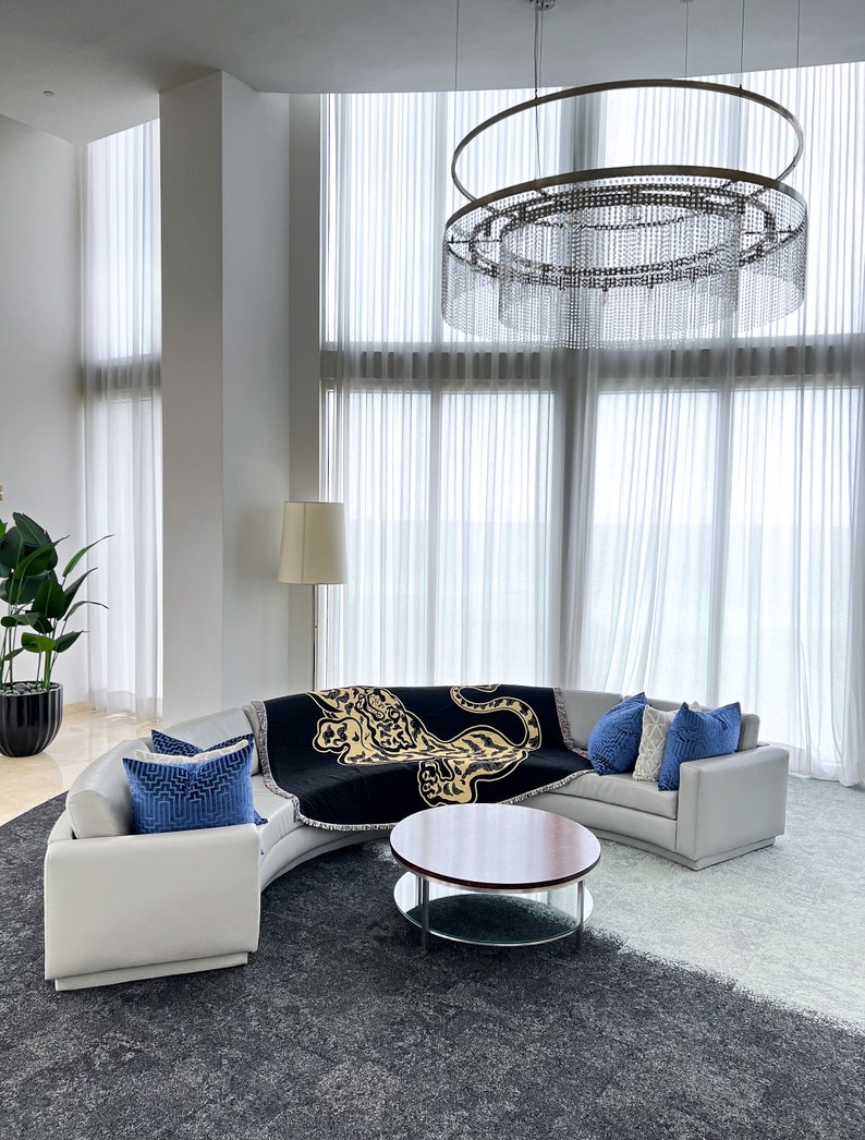May include: A white curved sectional sofa with blue throw pillows and a black throw blanket with a gold leopard print. The sofa is in a modern living room with a large crystal chandelier and a glass-top coffee table with a wooden top.