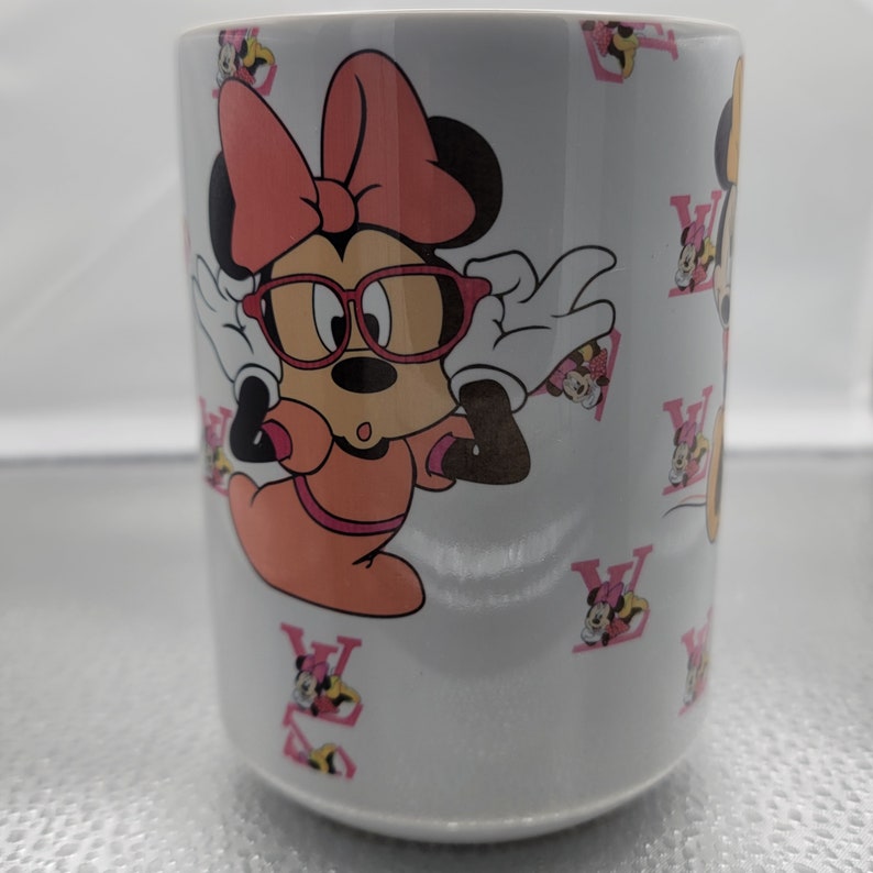 Pu&ograve; includere: Tazza in ceramica bianca con un design Minnie Mouse rosa e bianco. Minnie Mouse indossa occhiali rossi e un fiocco rosa. La tazza &egrave; decorata con un motivo ripetitivo di "V" rosa.