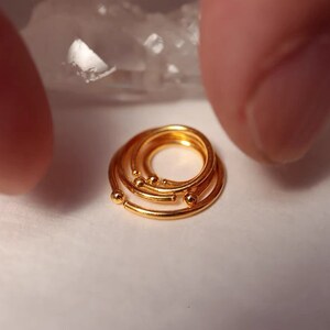 May include: A close-up of several small, gold-colored rings, each with a circular design. The rings have small, spherical accents. The jewelry is displayed on a white surface.