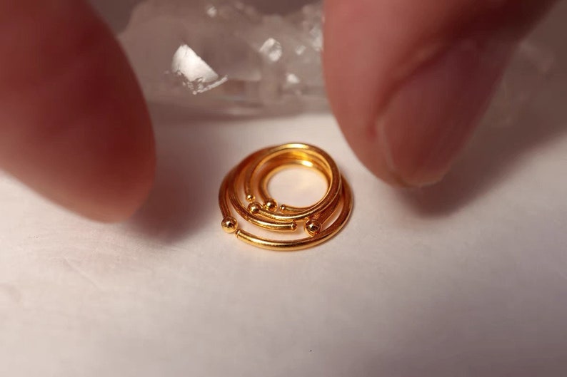May include: A close-up of several small, gold-colored rings, each with a circular design. The rings have small, spherical accents. The jewelry is displayed on a white surface.