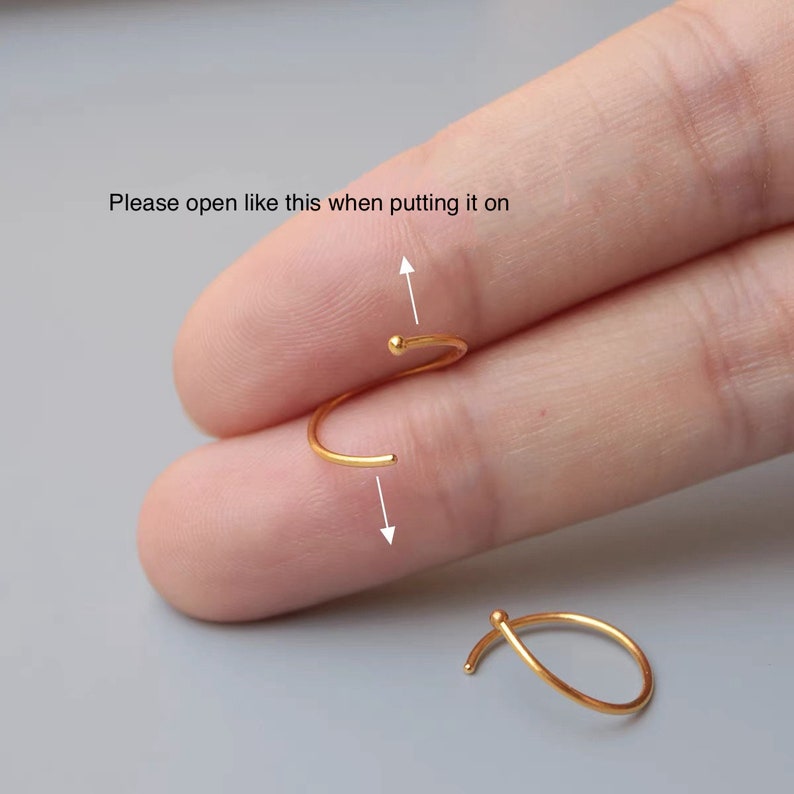 May include: A gold-colored earring with a curved design and a small ball at the end. The text in the image reads, "Please open like this when putting it on." The earring is shown being opened, with an arrow indicating the direction.