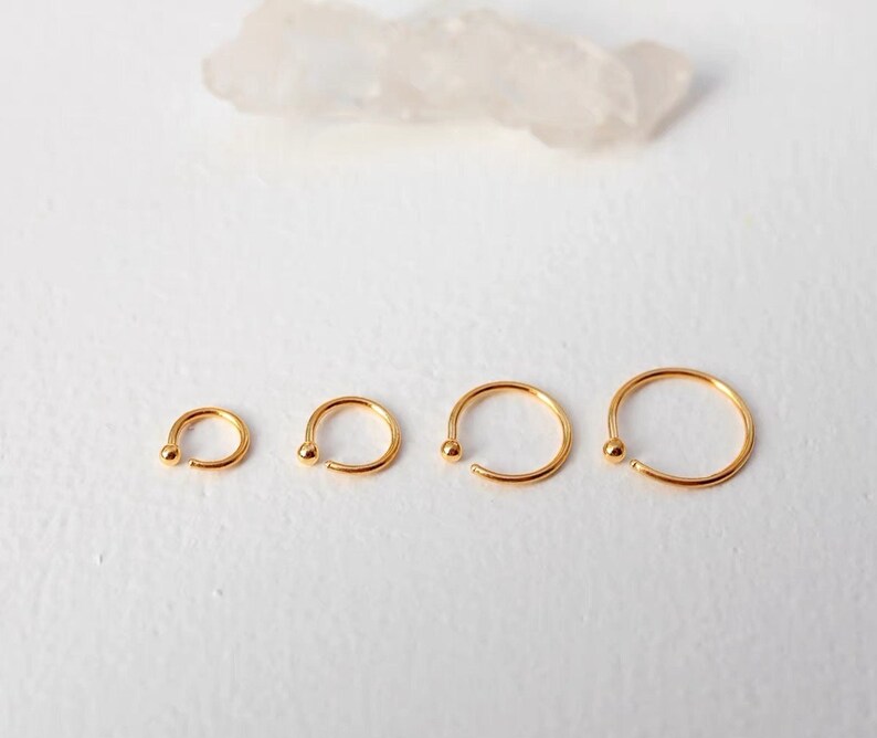 May include: Four gold-colored nose rings of varying sizes are displayed on a white surface. Each ring has a small ball at one end. The rings are arranged in a row, with the smallest on the left and the largest on the right.