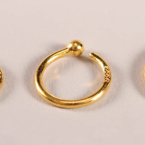May include: A close-up of a gold-colored ear cuff with a small ball at the top and the number "9999" engraved on the side. The ear cuff has a circular design and is set against a white background.