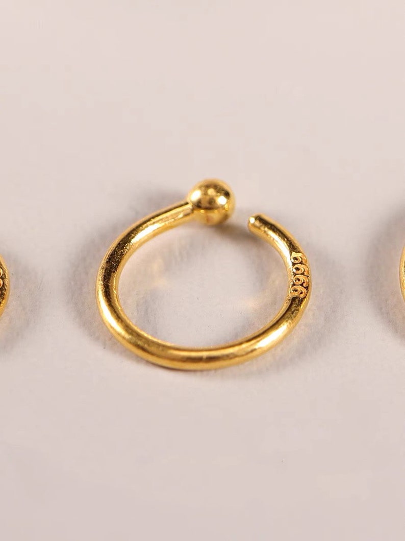 May include: A close-up of a gold-colored ear cuff with a small ball at the top and the number "9999" engraved on the side. The ear cuff has a circular design and is set against a white background.