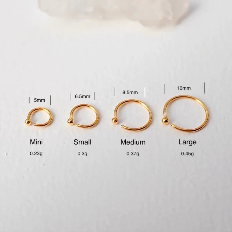 May include: Four gold-colored nose rings of varying sizes are displayed. The smallest is 5mm, labeled "Mini", and the largest is 10mm, labeled "Large". Each ring has a small ball at one end. The weights are also listed.