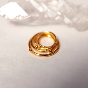 May include: A stack of gold-colored rings with small ball accents. The rings are arranged in a spiral, with a bright, reflective surface. The jewelry is set against a soft, white background.