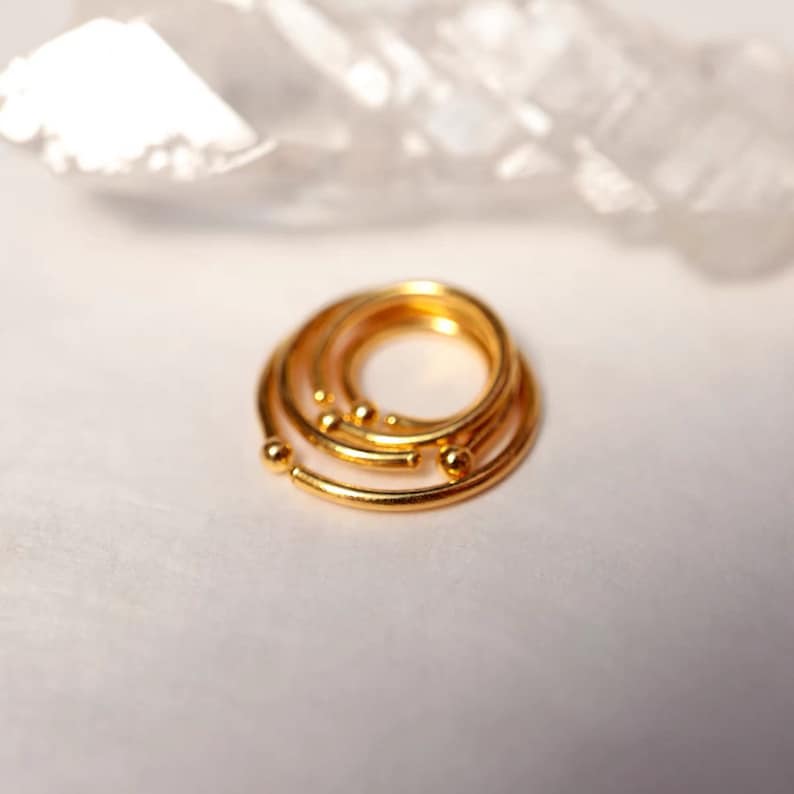 May include: A stack of gold-colored rings with small ball accents. The rings are arranged in a spiral, with a bright, reflective surface. The jewelry is set against a soft, white background.