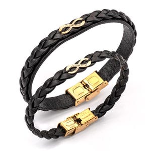 Op de afbeelding: Twee zwarte, gevlochten leren armbanden. Elke armband heeft een goudkleurig oneindigheidssymbool en een goudkleurige sluiting. De armbanden worden getoond tegen een witte achtergrond.