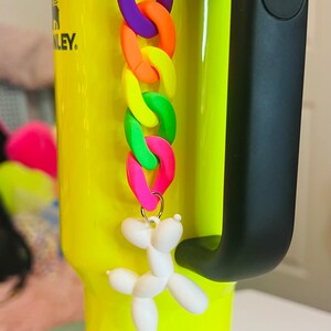 May include: A bright yellow travel mug with a black handle and a leopard print band around the bottom. A white balloon dog keychain with a rainbow chain is attached to the mug.
