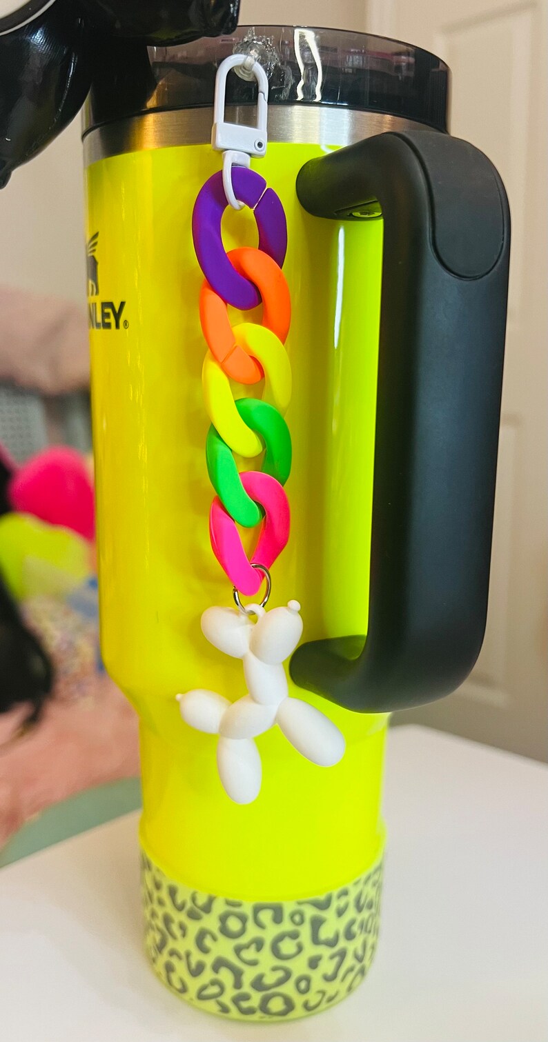 May include: A bright yellow travel mug with a black handle and a leopard print band around the bottom. A white balloon dog keychain with a rainbow chain is attached to the mug.