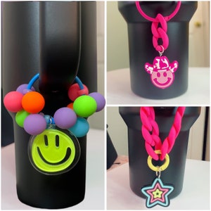 May include: Three black tumblers with different colorful charms. The first tumbler has a multi-colored beaded charm with a yellow smiley face. The second tumbler has a pink cowboy hat charm with a smiley face. The third tumbler has a pink chain charm with a yellow star.