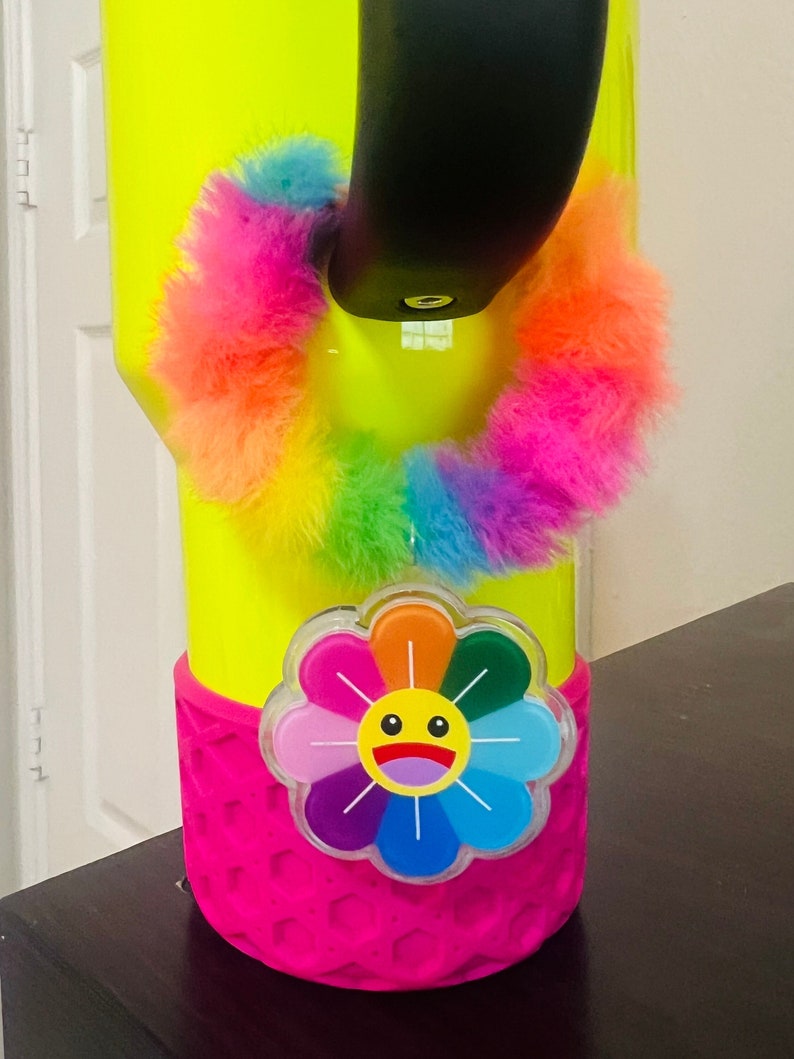May include: A neon yellow tumbler with a pink silicone base and a rainbow fuzzy ring. A colorful flower with a smiley face is attached to the base.