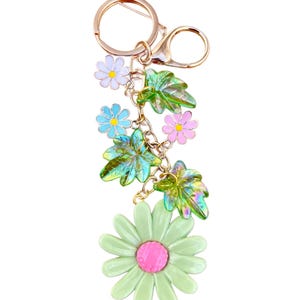 May include: A floral-themed keychain with a gold-tone ring and clasp. The keychain features a large green daisy with a pink center, along with smaller blue, pink, and white flowers, and iridescent green leaves. The design is connected by a gold chain.