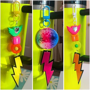 May include: Three colorful keychains with different charms. The first keychain has a rainbow disco ball charm, a pink lightning bolt charm, and a heart charm with a blue gem. The second keychain has a yellow lightning bolt charm, a red and white block charm, and a yellow and orange circle charm. The third keychain has an orange lightning bolt charm, a pink and white block charm, and a green and white circle charm.