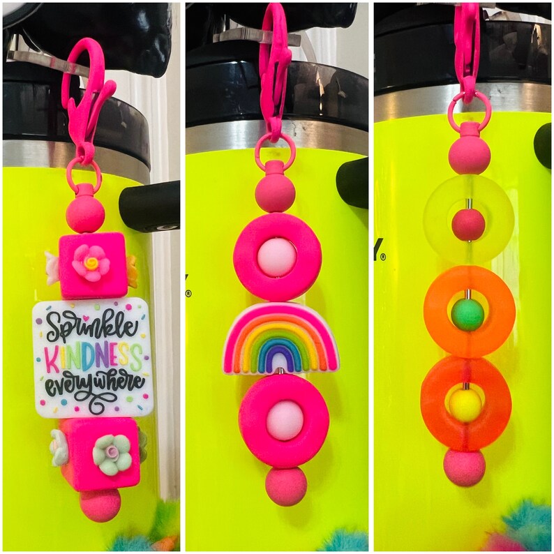 May include: Three colorful silicone charms with different designs. The first charm has a pink square with a flower and a white square with the text "Sprinkle Kindness Everywhere". The second charm has a pink circle with a white circle inside, a rainbow, and another pink circle. The third charm has three pink circles with a smaller circle inside each.