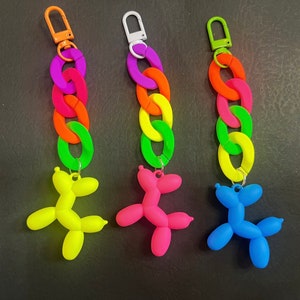 May include: Three colorful keychain charms featuring a rainbow chain and a balloon animal shaped like a dog. The balloon animals are yellow, pink, and blue.