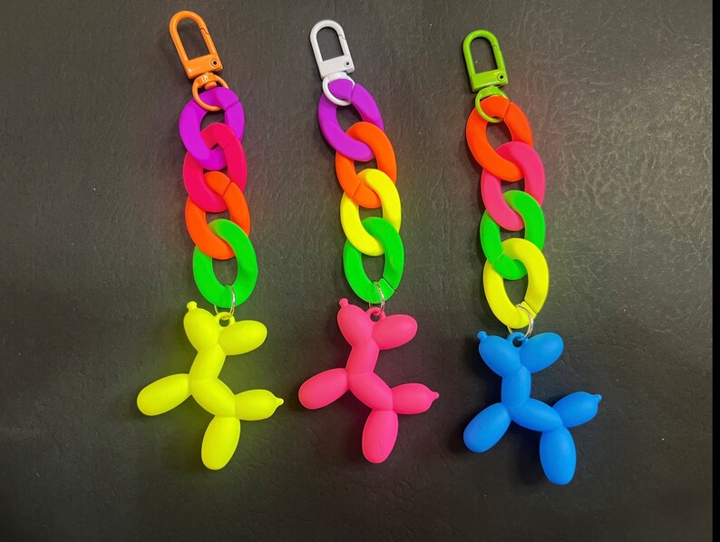 May include: Three colorful keychain charms featuring a rainbow chain and a balloon animal shaped like a dog. The balloon animals are yellow, pink, and blue.