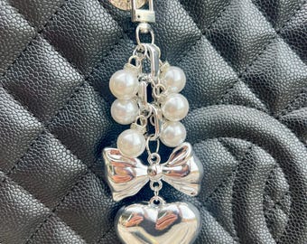 May include: Silver-toned bag charm with a heart pendant, bow, and faux pearl accents. The charm is attached to a silver ring and clasp. The background is a black, quilted leather handbag.