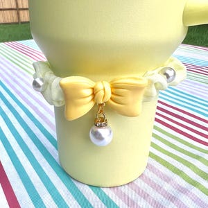 May include: A yellow ceramic vase with a white and yellow bow and a pearl pendant. The vase is sitting on a striped tablecloth.
