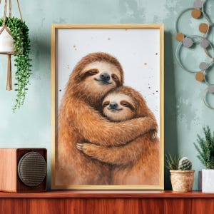 May include: A framed watercolor print featuring two sloths embracing. The sloths are depicted with brown fur and smiling faces. The artwork has a warm, inviting feel, with a light background and subtle paint splatters. The frame is a light wood color.