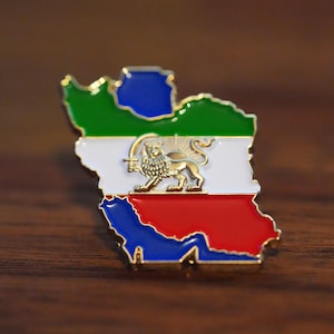 Puede incluir: Un colorido pin de esmalte con forma de mapa, con secciones verdes, blancas, rojas y azules. Un emblema de león dorado está centrado en la parte blanca. El pin está sobre una superficie de madera oscura.