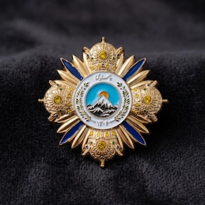Könnte beinhalten: Ein aufwendiges, goldfarbenes Emblem mit einem zentralen Design, das eine Bergkette, eine aufgehende Sonne und Text in einem kreisförmigen Rahmen zeigt. Das Emblem hat blaue und weiße Emaille-Akzente und ist vor einem dunklen Hintergrund platziert.