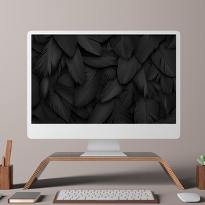 Op de afbeelding: Een witte computermonitor toont een close-up van zwarte veren. De monitor staat op een houten standaard, met een toetsenbord en muis ervoor. Een houten potloodhouder en een klein notitieboekje staan links.