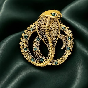 May include: A gold-toned metal brooch shaped like a coiled cobra snake. The textured surface of the snake features a series of small, round, dark green rhinestones embedded along its body and a single green stone set as the eye of the cobra.