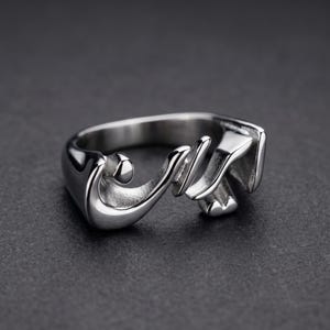 Puede incluir: Un anillo plateado con un diseño en cursiva. El anillo presenta un patrón fluido en forma de guion, con un remolino redondeado al principio y un extremo estilizado. El anillo está sobre un fondo gris oscuro.