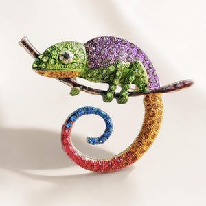 May include: A brooch shaped like a chameleon perched on a branch. The chameleon is covered in multicoloured rhinestones, featuring a green head, purple back, and a long, spiraled tail that transitions from blue to red and orange hues.