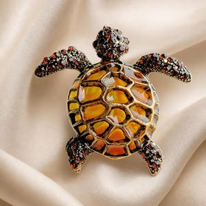 Könnte beinhalten: Eine schildkrötenförmige Brosche mit einem Panzer aus durchscheinenden bernsteinfarbenen und orangefarbenen Emaille-Segmenten, die mit goldfarbenem Metall eingefasst sind. Kopf und Flossen sind mit kleinen, braunen, facettierten Steinen besetzt.