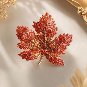 Könnte beinhalten: Eine Ahornblatt-Brosche mit einem goldfarbenen Metallrahmen, der mit zahlreichen kleinen, leuchtend orangefarbenen und roten Kristallen besetzt ist. Das Design enthält mehrere größere, runde, facettierte orangefarbene Steine.