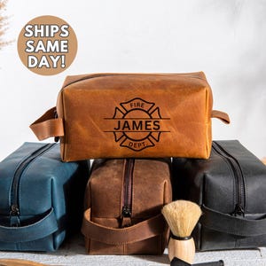 May include: A collection of leather toiletry bags in various colors, including brown, blue, and black. The top bag is embossed with "FIRE JAMES DEPT." and a fire department emblem. Each bag has a zipper closure and a handle.