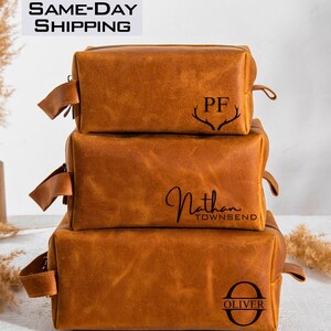 May include: Three brown leather toiletry bags stacked on top of each other. The top bag has the initials "PF" with antlers above the letters. The middle bag has the name "Nathan Townsend" written in cursive. The bottom bag has the letter "O" with the name "Oliver" written below it.