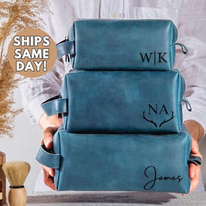 May include: Three blue leather toiletry bags with different personalized monograms. The top bag has the initials "W|K", the middle bag has the initials "NA" with antlers, and the bottom bag has the name "James" in cursive.